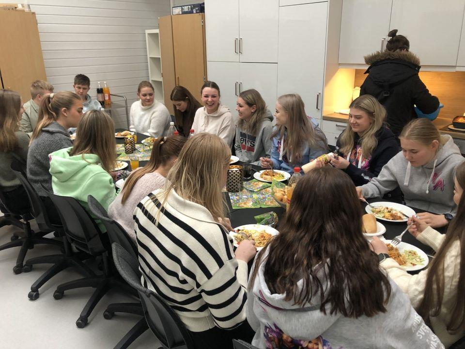 Mange elever sitter rundt et bord og spiser lasagne. - Klikk for stort bilde